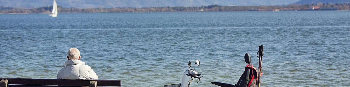 Back view of older man sitting on a bench on the waterfront with an electric wheelchair by his side.
